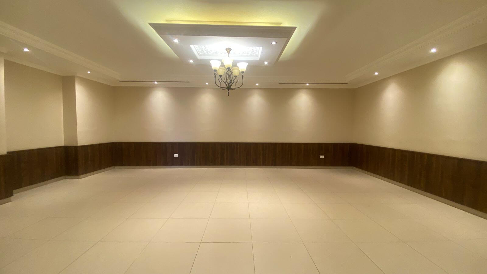 869c197f-7a76-4a8c-b4a3-51721d9e2a91 Empty banquet hall with cream walls, dark wood wainscoting, and a tiled floor under a chandelier.
