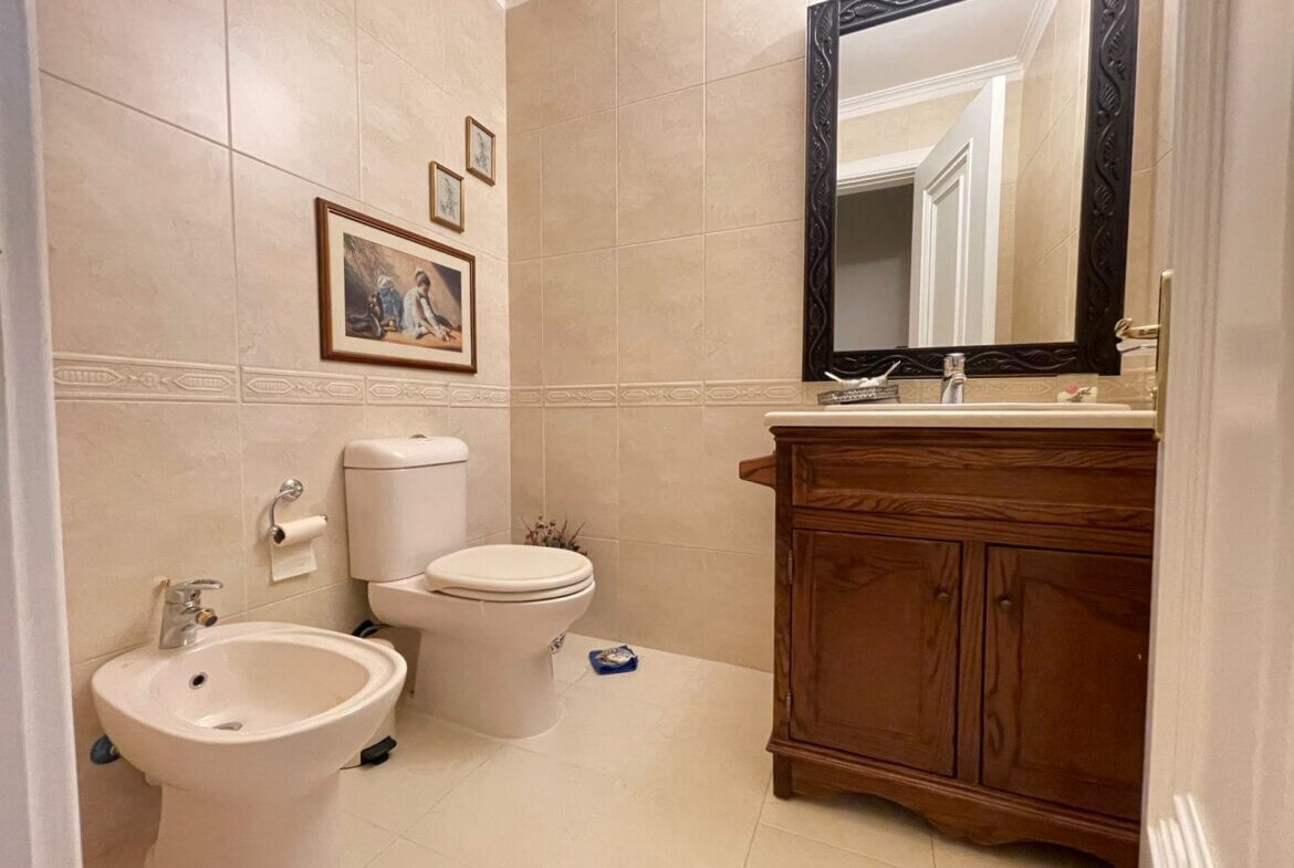 87098436-37b2-40a6-b174-1c555251cb79 Beige-tiled bathroom with a wooden vanity, large ornate black-framed mirror, and a white toilet and bidet.