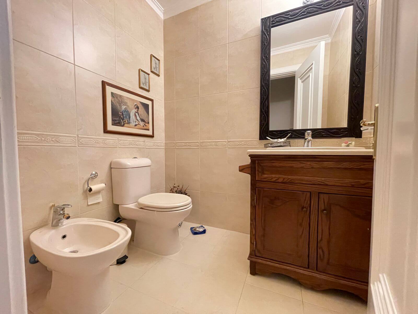 87098436-37b2-40a6-b174-1c555251cb79 Beige-tiled bathroom with a wooden vanity, large ornate black-framed mirror, and a white toilet and bidet.