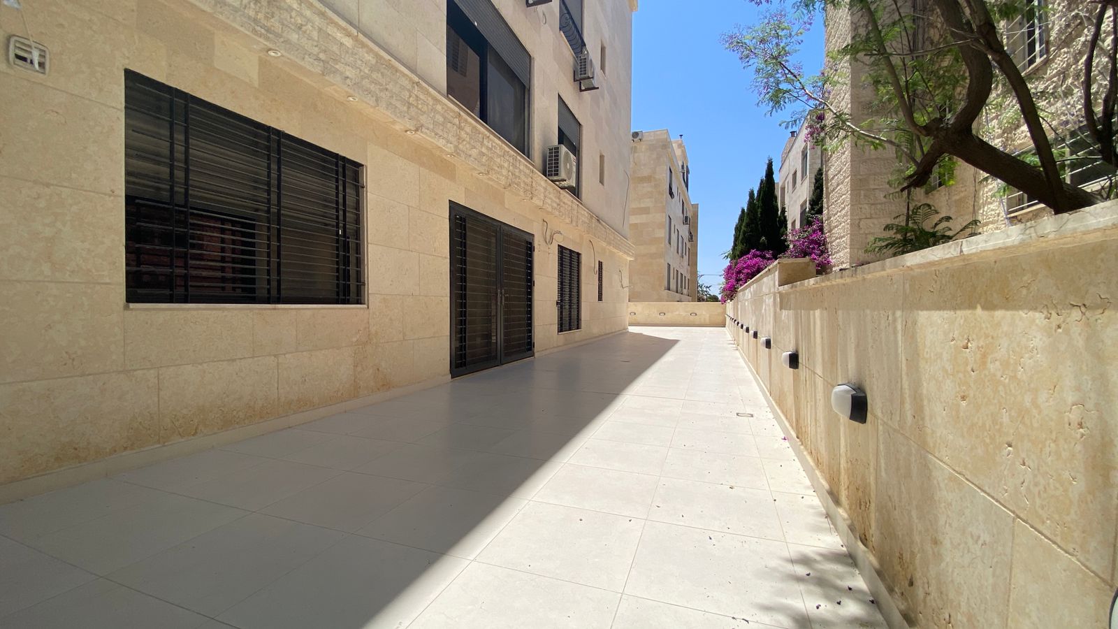 8759a0ab-964a-4a85-98e3-d91d299de0e5 Sunny beige stone alley between apartment buildings with barred windows, white tile walkway, and a long shadow across the ground, blue sky above.