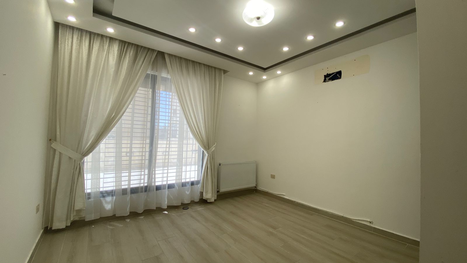 876e1d86-2476-4088-ad64-8054bc0665f2 Empty modern living room with large window covered by sheer white curtains, light wood flooring, and a row of recessed ceiling lights.