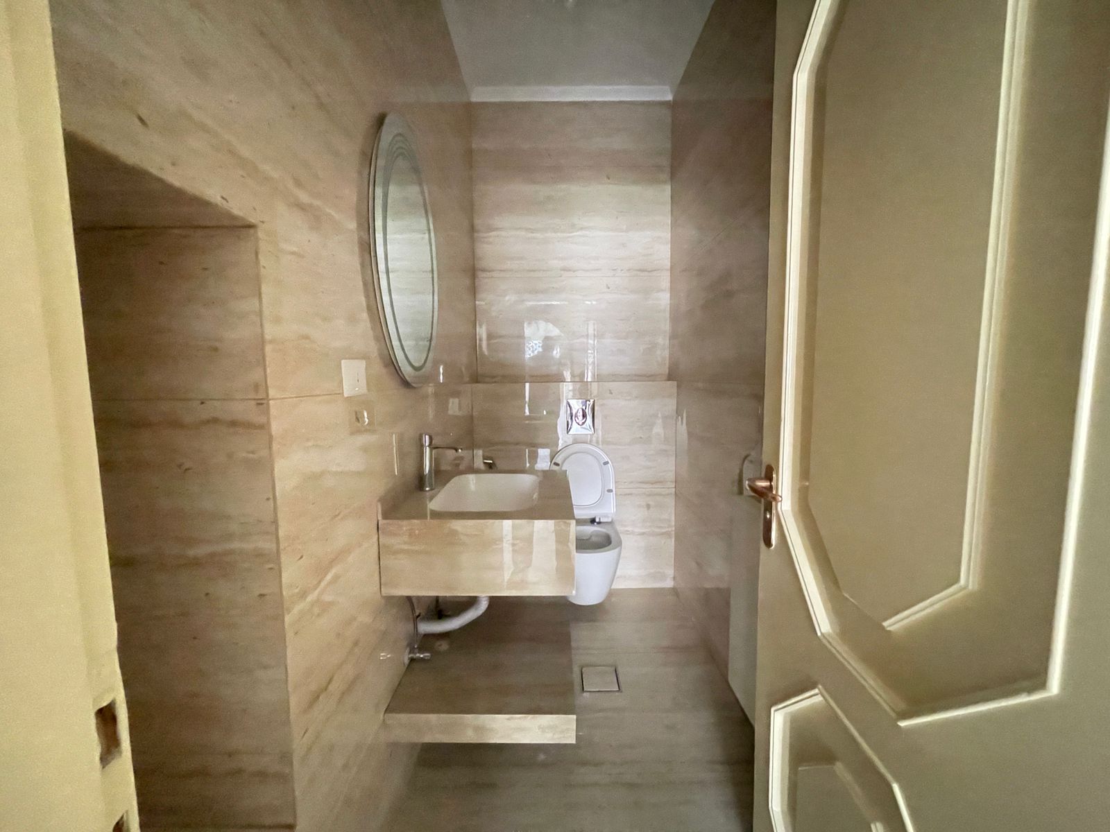 880077e8-9aa8-45fe-9564-6164b1d5435a Beige marble bathroom showing a wall-mounted sink, round mirror, and a toilet with a wooden door to the right.