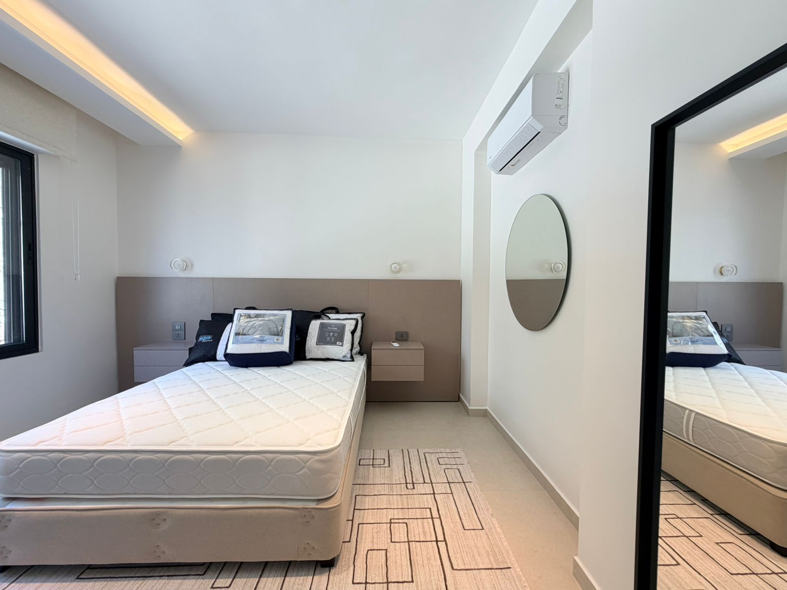 894fa8d7-e9c1-466e-bcc3-b5de0aa2d75d Minimalist hotel bedroom with a white-tiled mattress on a low bed frame, beige headboard, two floating nightstands, and a large circular mirror on the opposite wall to the right