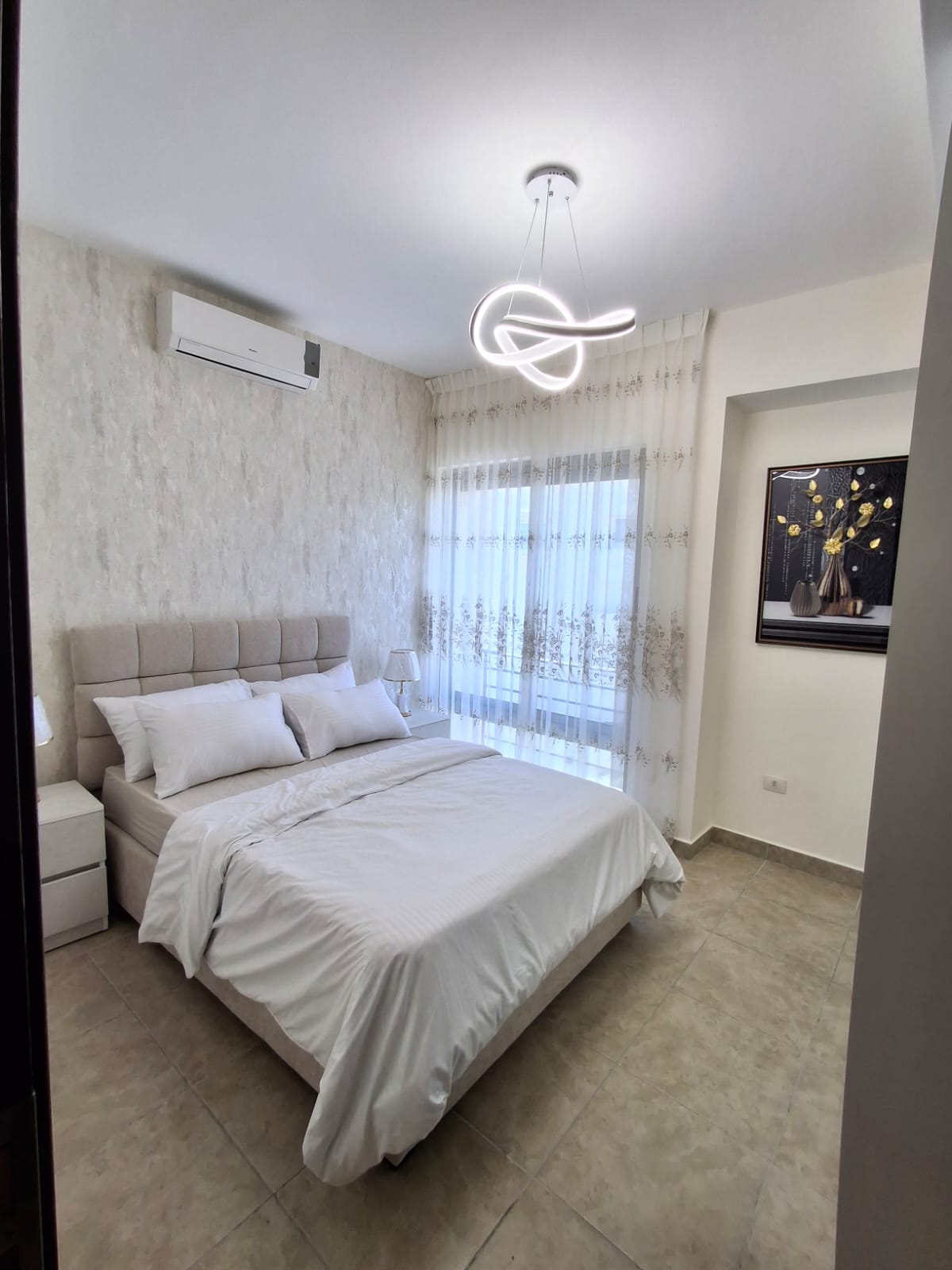 89630705-8ee9-4ffd-84fb-cca6e0913d41 Modern bedroom with white bedding and a tufted beige headboard, sheer patterned curtains, and a sculptural loop chandelier above the bed