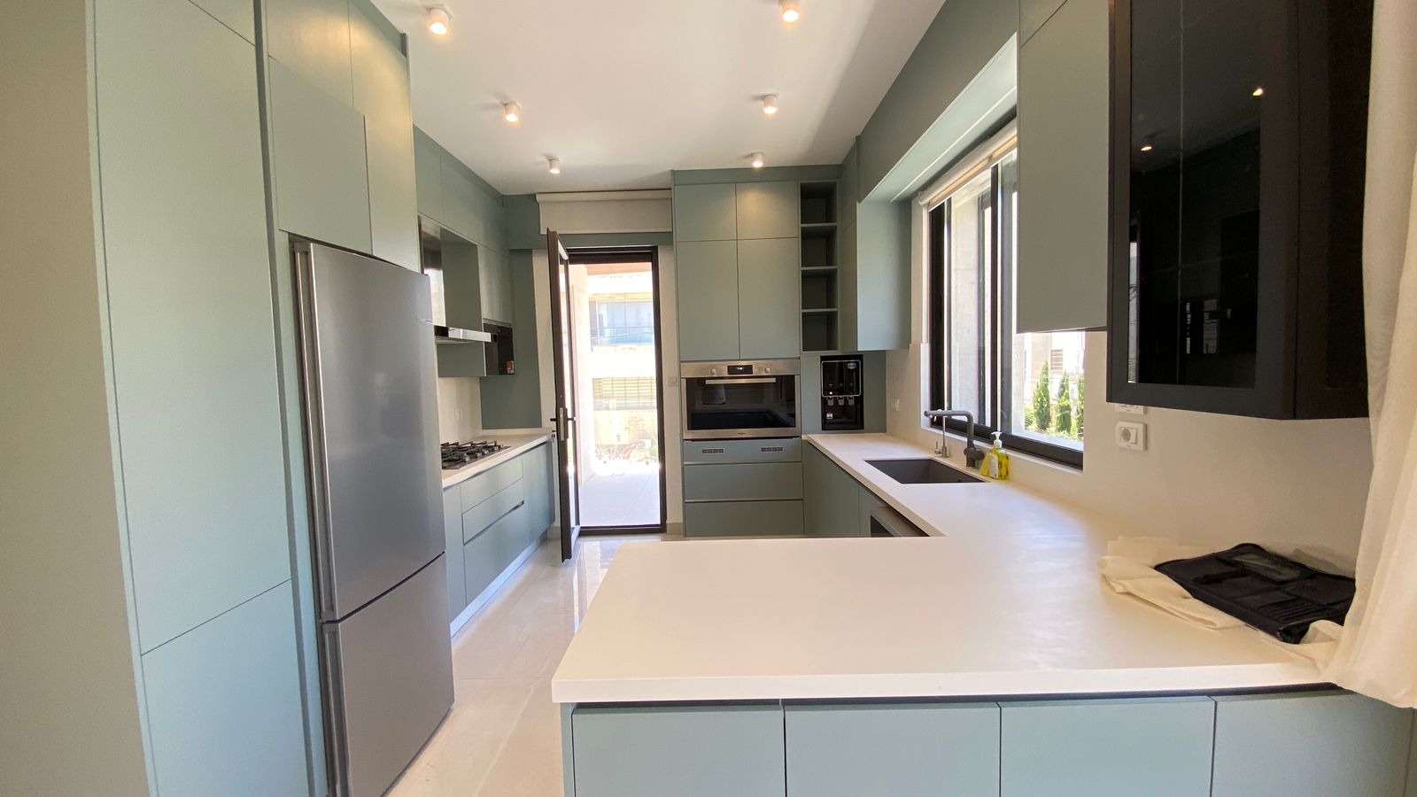 89d55118-ab1e-4c34-a0a3-8a04b1d8b4f1 Modern kitchen with pale green flat-front cabinets, stainless steel fridge, and a white countertop island. A bright window brings in daylight.