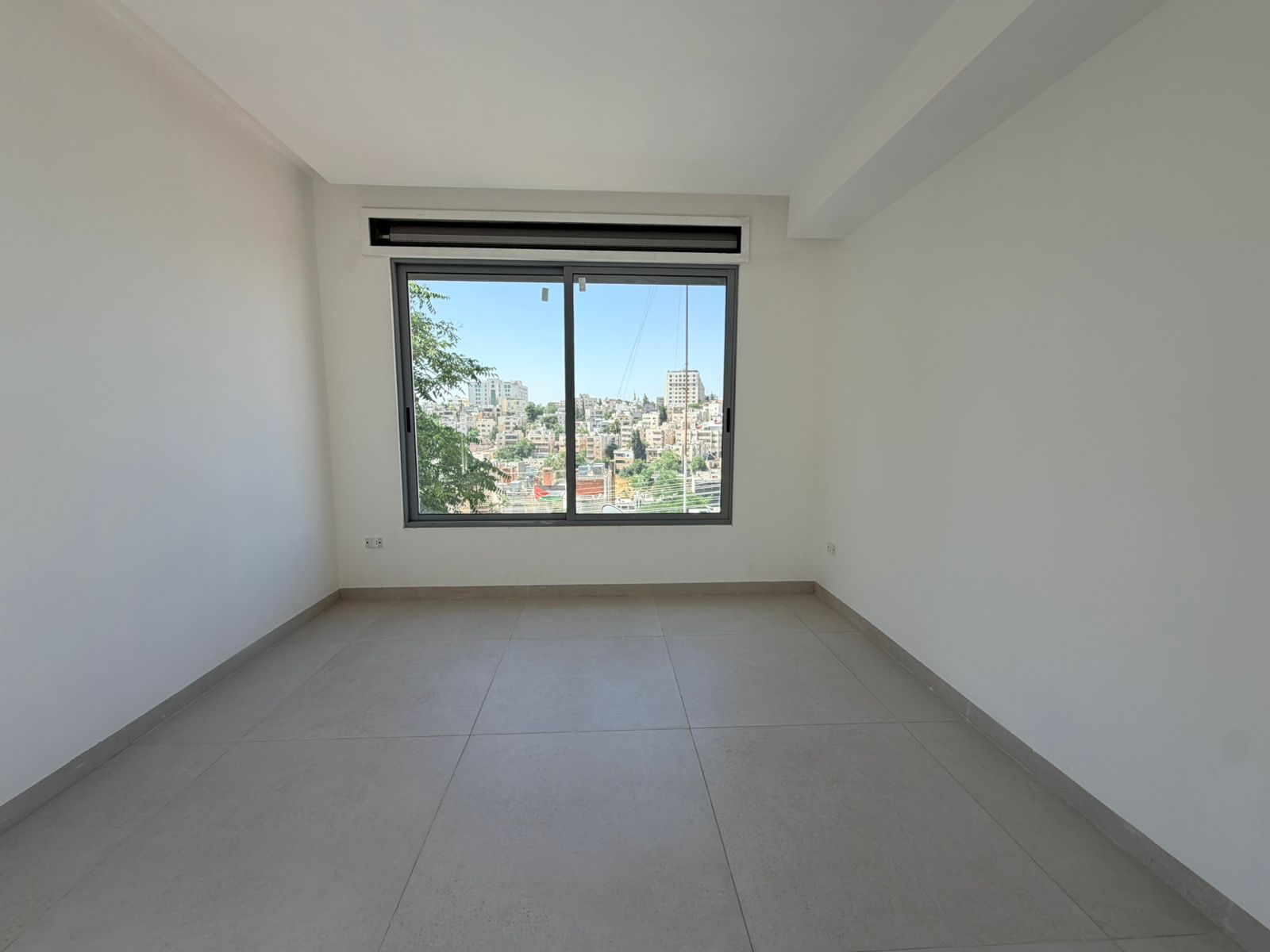 8a3d9fbf-e5d6-4004-be07-72b2a53546d8 Vacant white room with a large sliding window looking out on a cityscape and trees; light gray tiled floor and an overhead vent above the window.