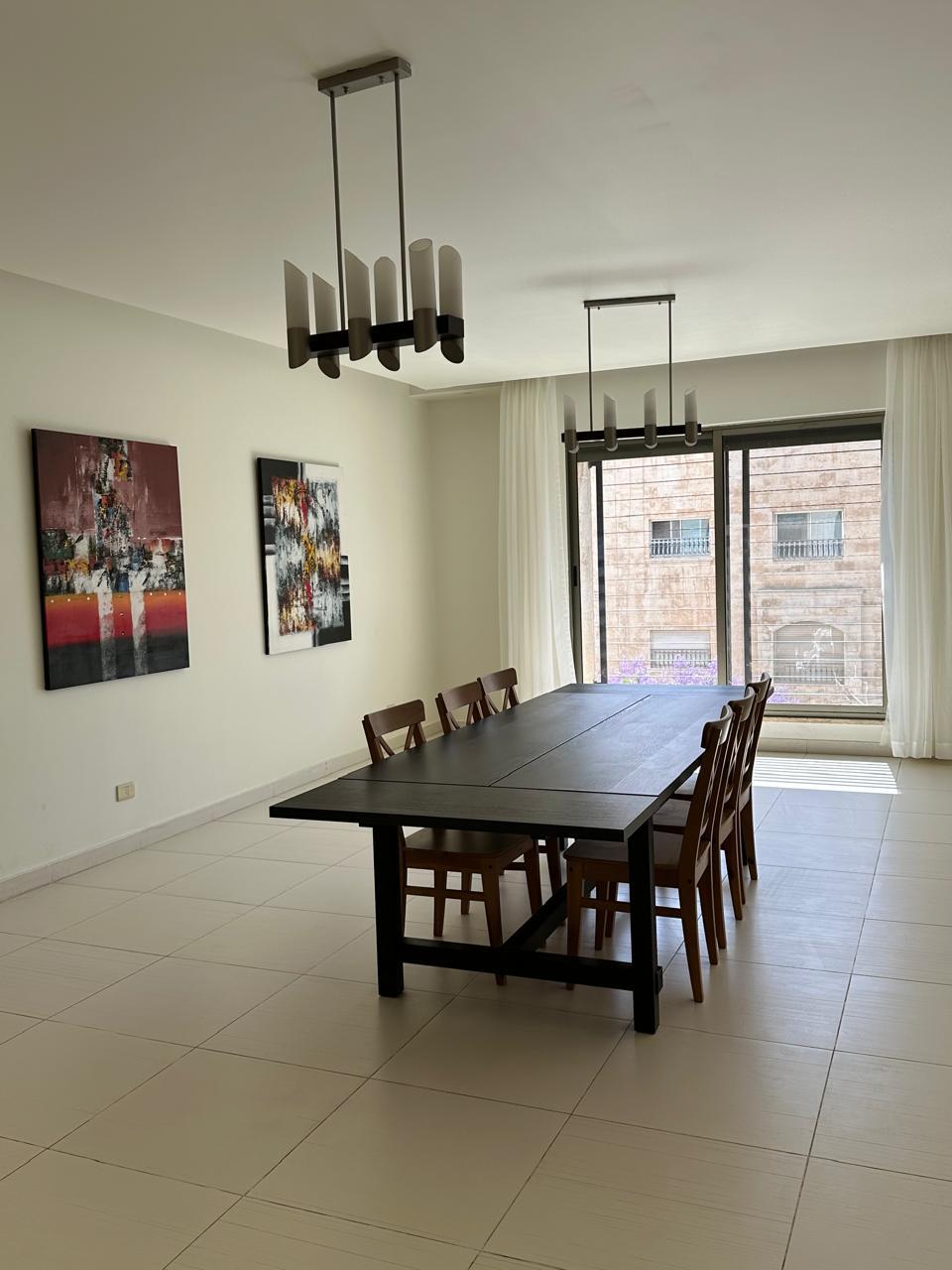 8b0fc933-ffef-4309-8602-5371153ad52c Long dark dining table with eight wooden chairs in a bright room, two modern pendant lights, and a large window with sheer white curtains.