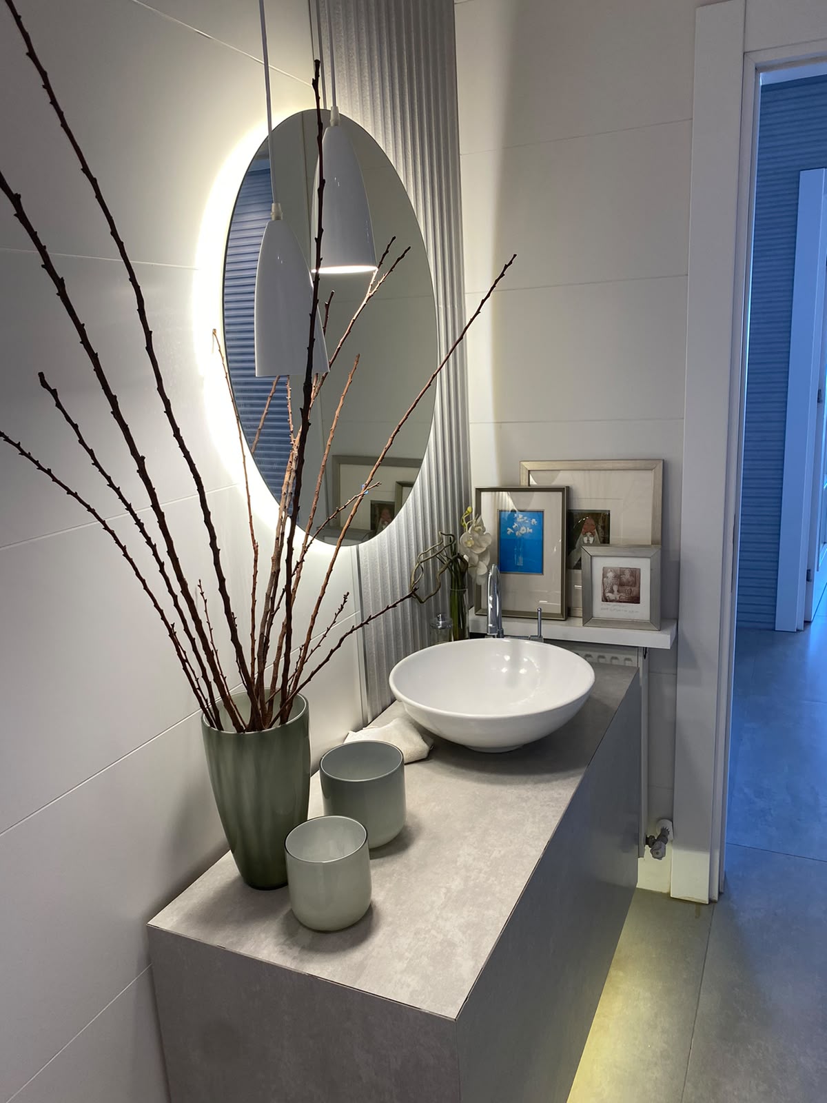 8b400dff-0207-485d-a51d-5f4d2ab38316 Modern bathroom vanity with a round backlit mirror, white vessel sink, and decorative vases with bare branches on a light countertop.