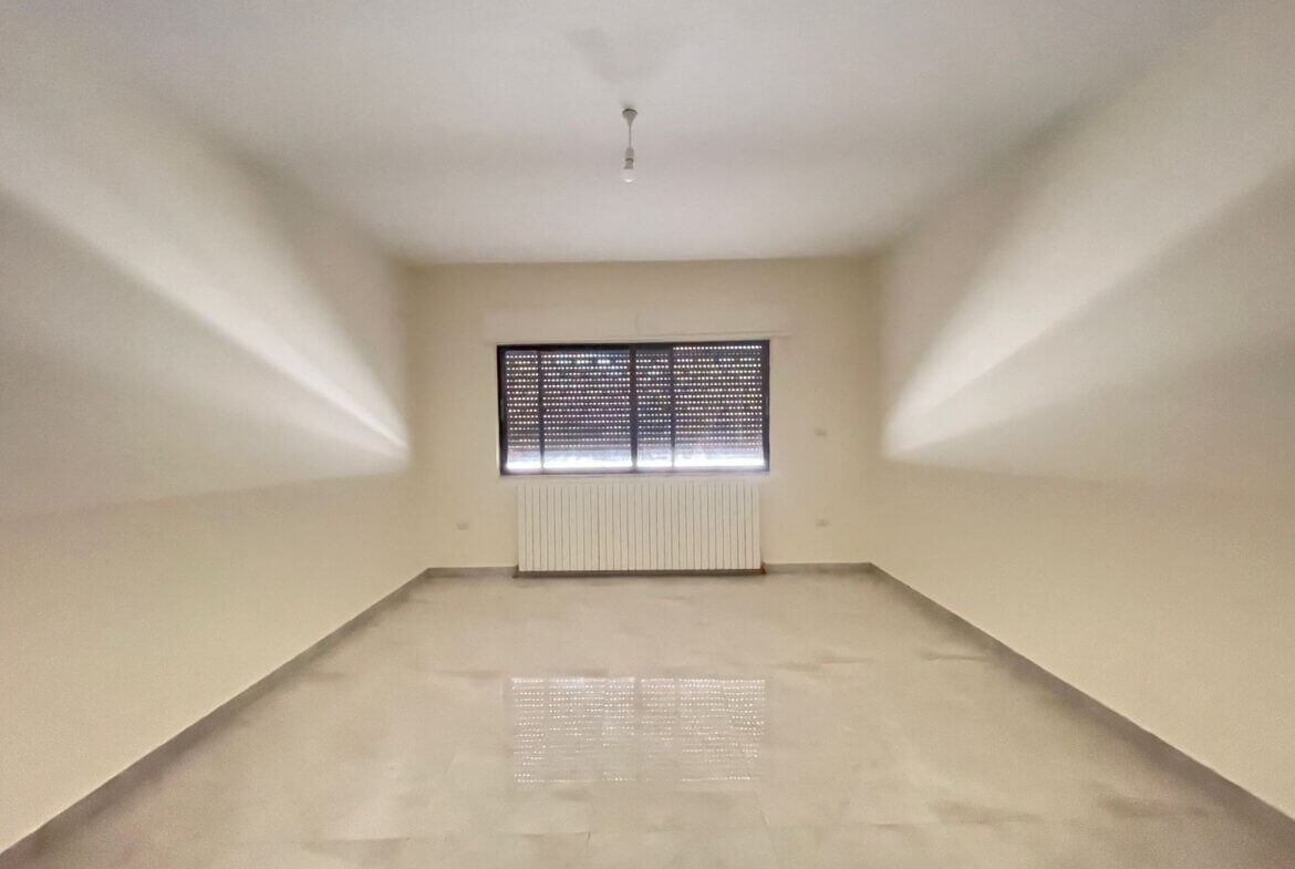 8b893b05-4024-4462-bab7-e055f18b3154 Empty rectangular room with pale walls, a window with shutters, radiator beneath, and a single ceiling light.