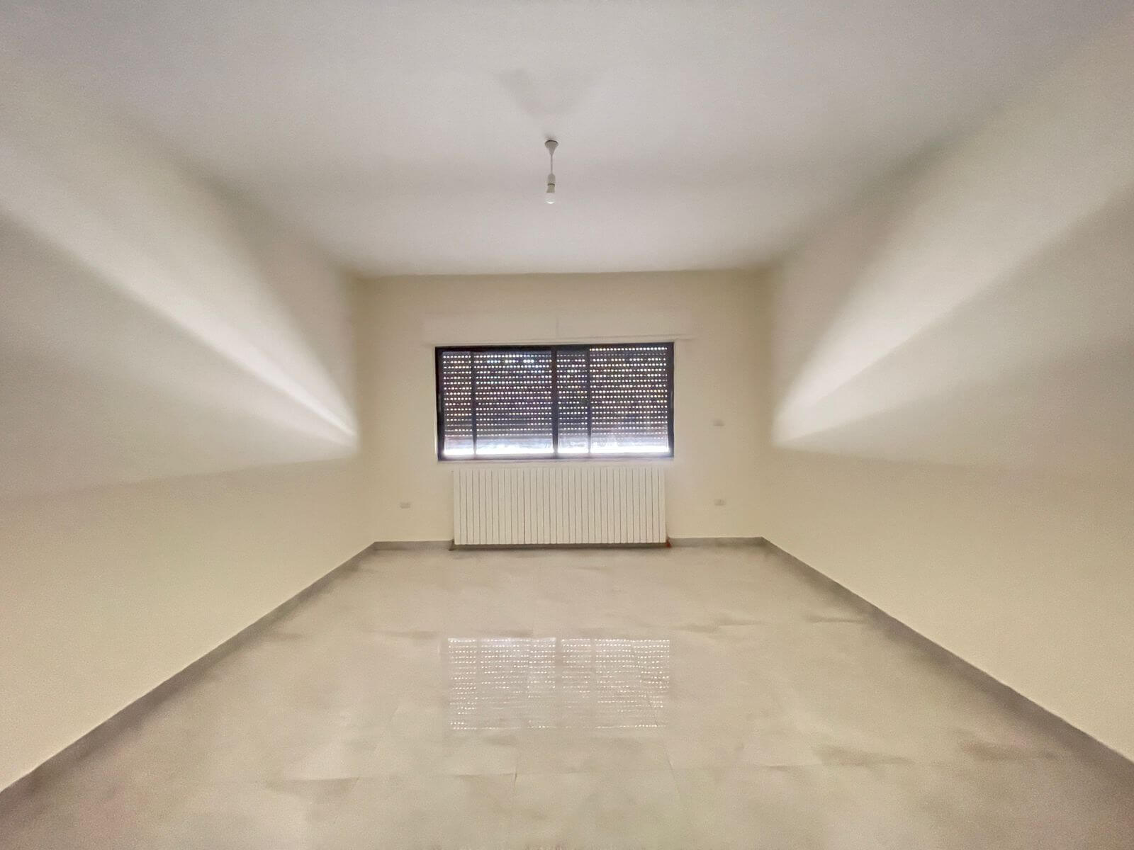 8b893b05-4024-4462-bab7-e055f18b3154 Empty rectangular room with pale walls, a window with shutters, radiator beneath, and a single ceiling light.