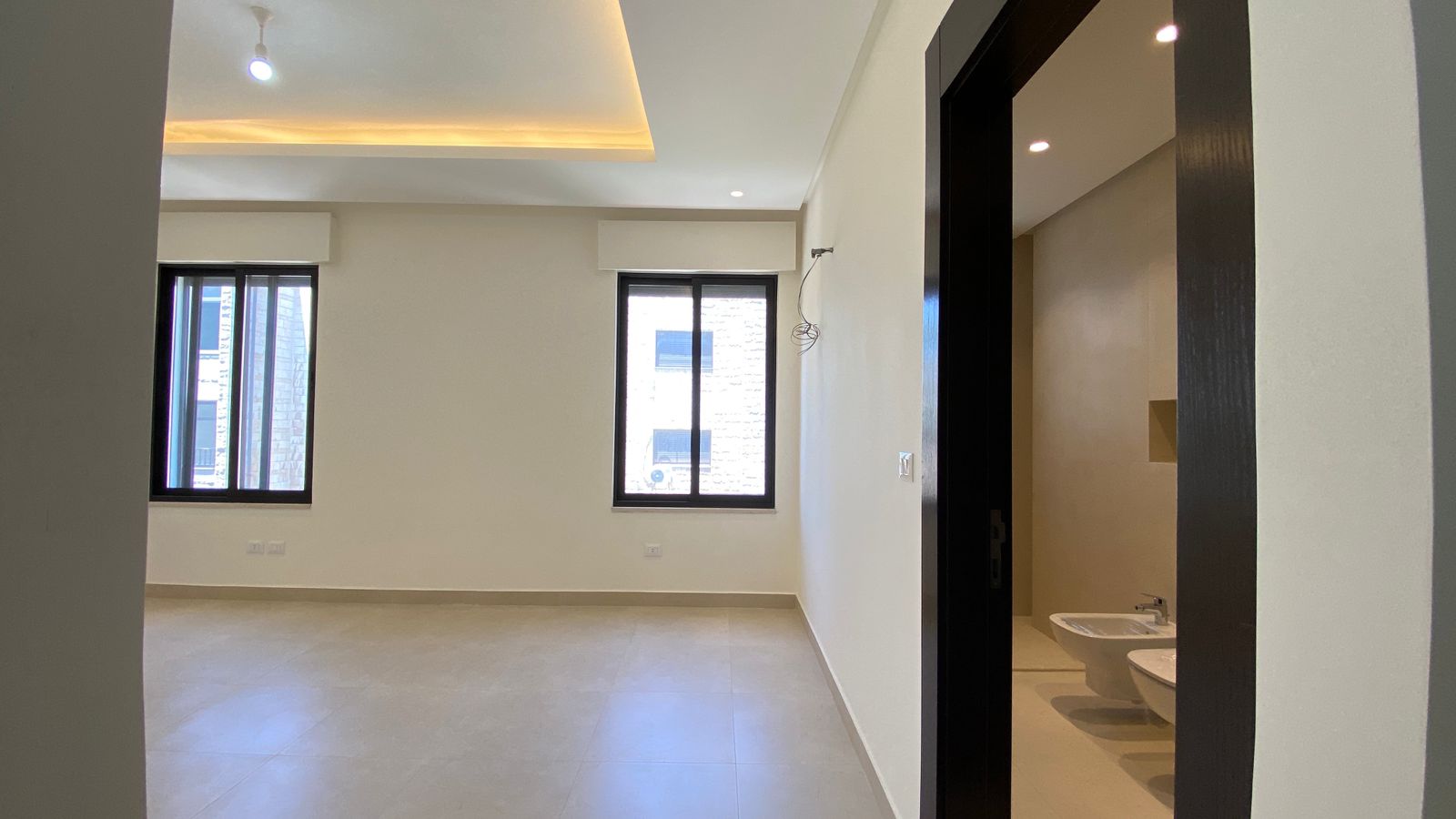 8c6f7b50-7002-4f03-a434-646ebdec4fc6 Unfurnished living room in a modern apartment with two windows, beige walls, and a doorway to a bathroom showing a toilet and bidet; ceiling has recessed lighting and exposed wiring.