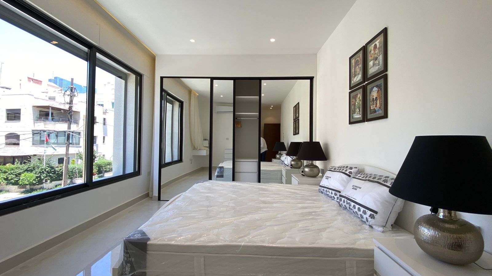 8ec2e7aa-bec3-48d9-8824-6d9f13346286 Bright, modern bedroom with large windows on the left, a white bed in the foreground, and a mirrored sliding-door wardrobe at the far end, plus bedside lamps and framed wall art.