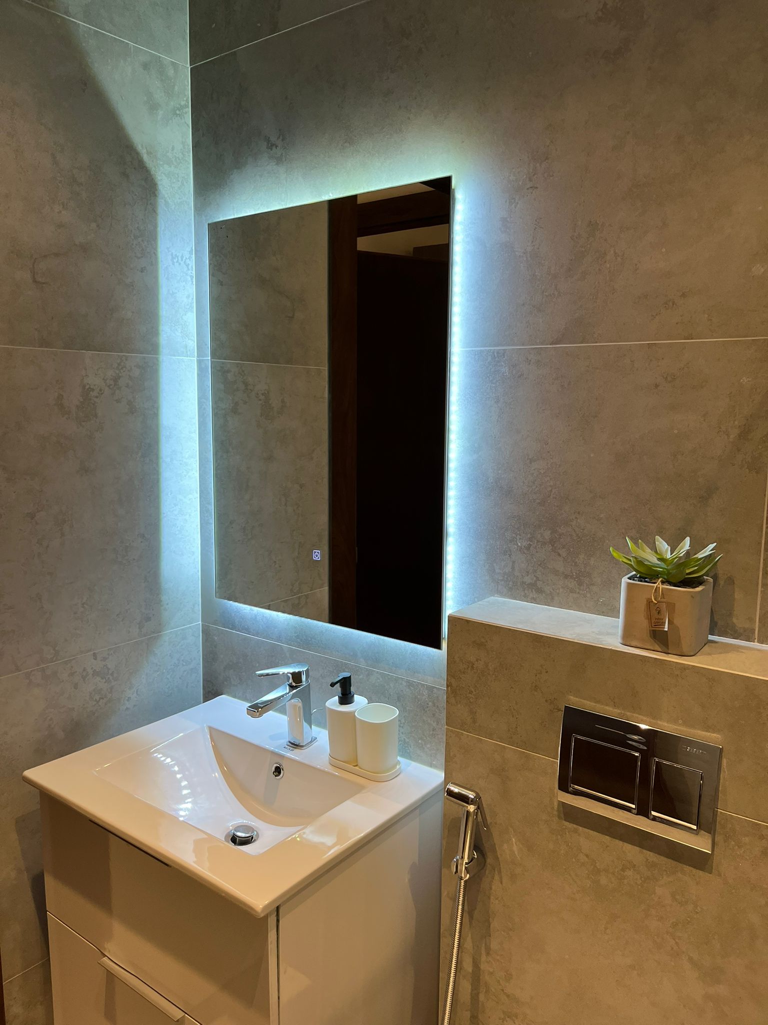 8f676b7c-ee26-481d-b64c-432de875da23 Modern bathroom vanity with white sink, backlit rectangular mirror, and gray tiled walls; a small plant on the shelf nearby.