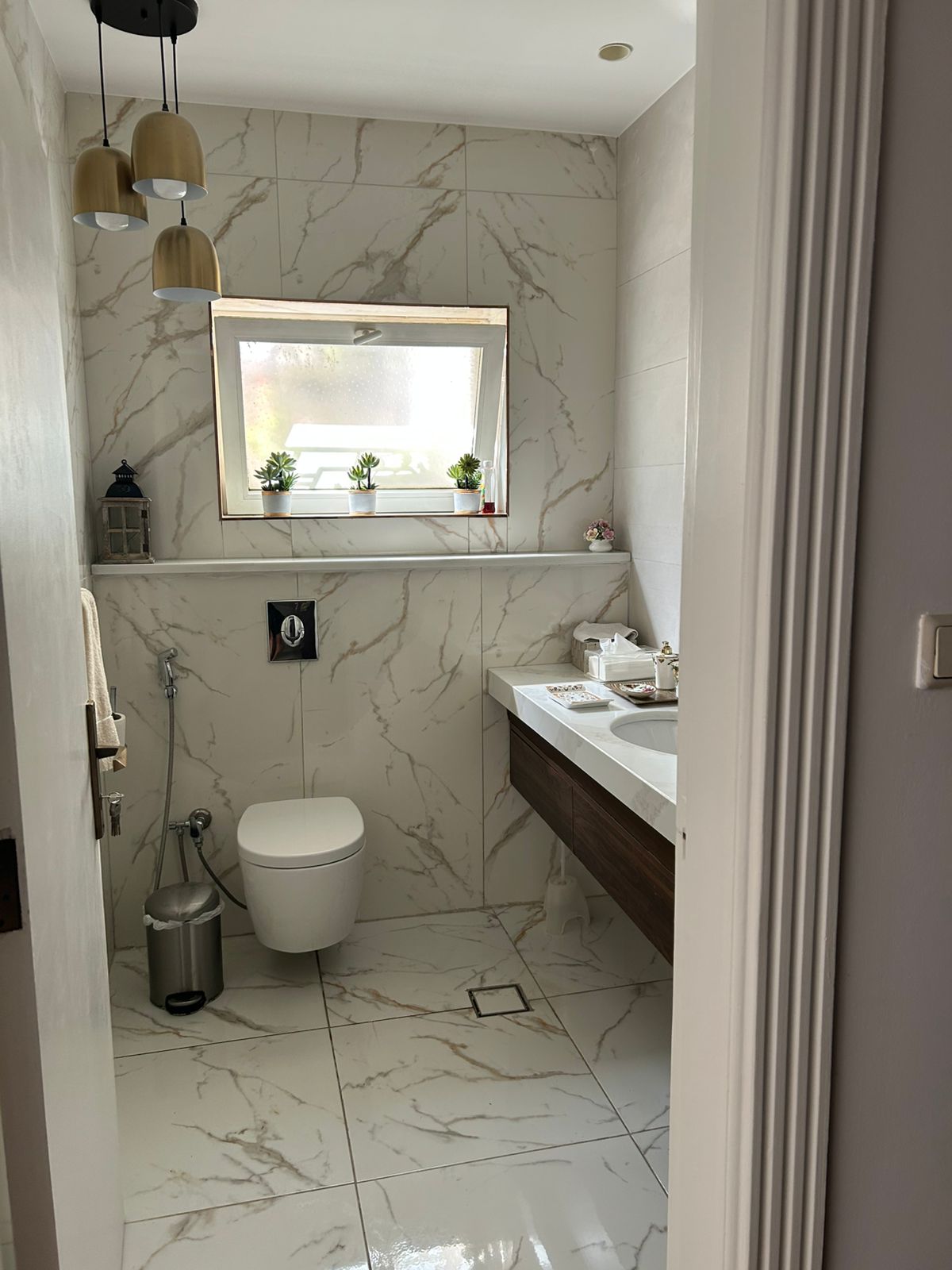9052bed7-2290-491d-b7a8-c8d41ba832af Modern marble-tiled bathroom with a wall-hung toilet, white sink, and a rectangular window ledge with small potted plants.