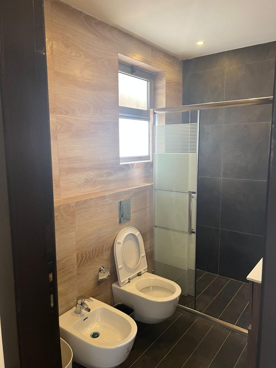 90571e82-5400-464b-ba0a-52ae39b53336 Modern bathroom with wood-paneled walls, a frosted-glass shower enclosure, toilet and bidet, and a small window above. Dark floor tiles complete the contemporary look.