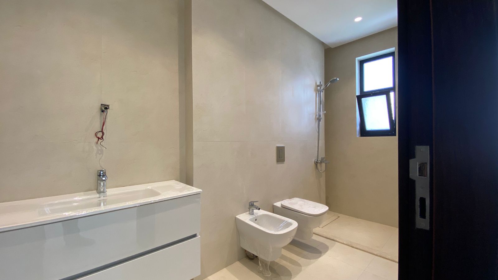 9062ee13-a2a9-4401-9776-1349d42fc9a7 Modern beige-tiled bathroom with a wall-mounted vanity and sink on the left, exposed wiring above, and a bidet and wall-hung toilet in the center; a shower area with a handheld head is at the far end by a window.