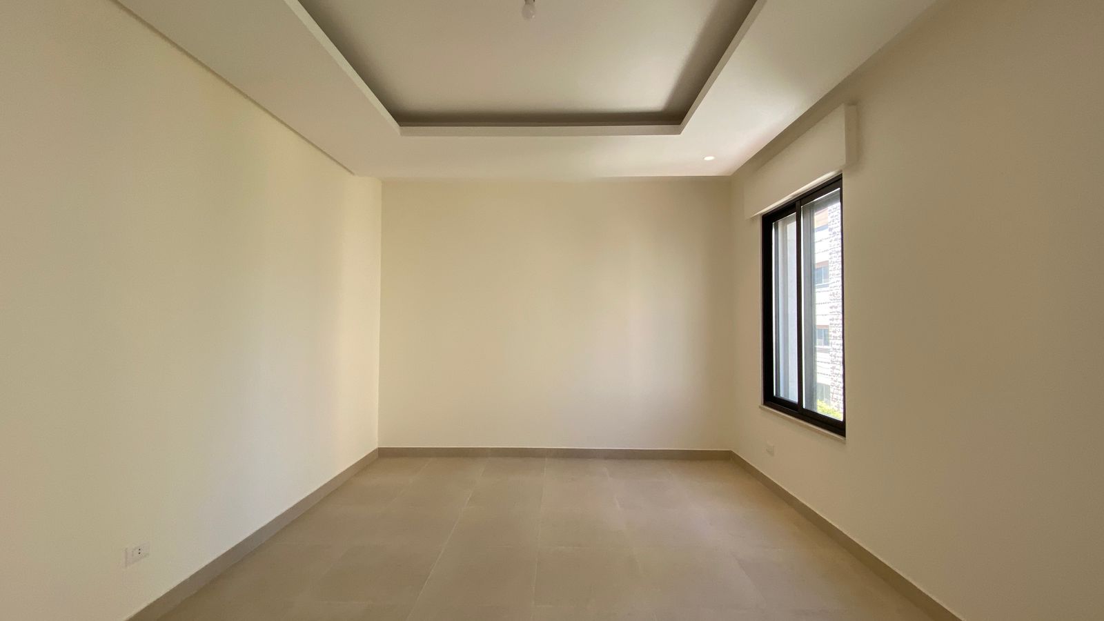 9080a554-8304-4e7b-8583-5d3535c27920 Empty modern room with beige walls, light tiled floor, and a large window on the right; recessed ceiling feature adds depth.