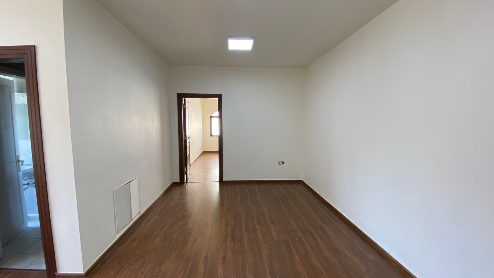 90edc40b-58f3-4760-92a3-005579e90a5d Empty living room with dark wood floors, white walls, a ceiling light, and a doorway to a sunlit adjacent room.