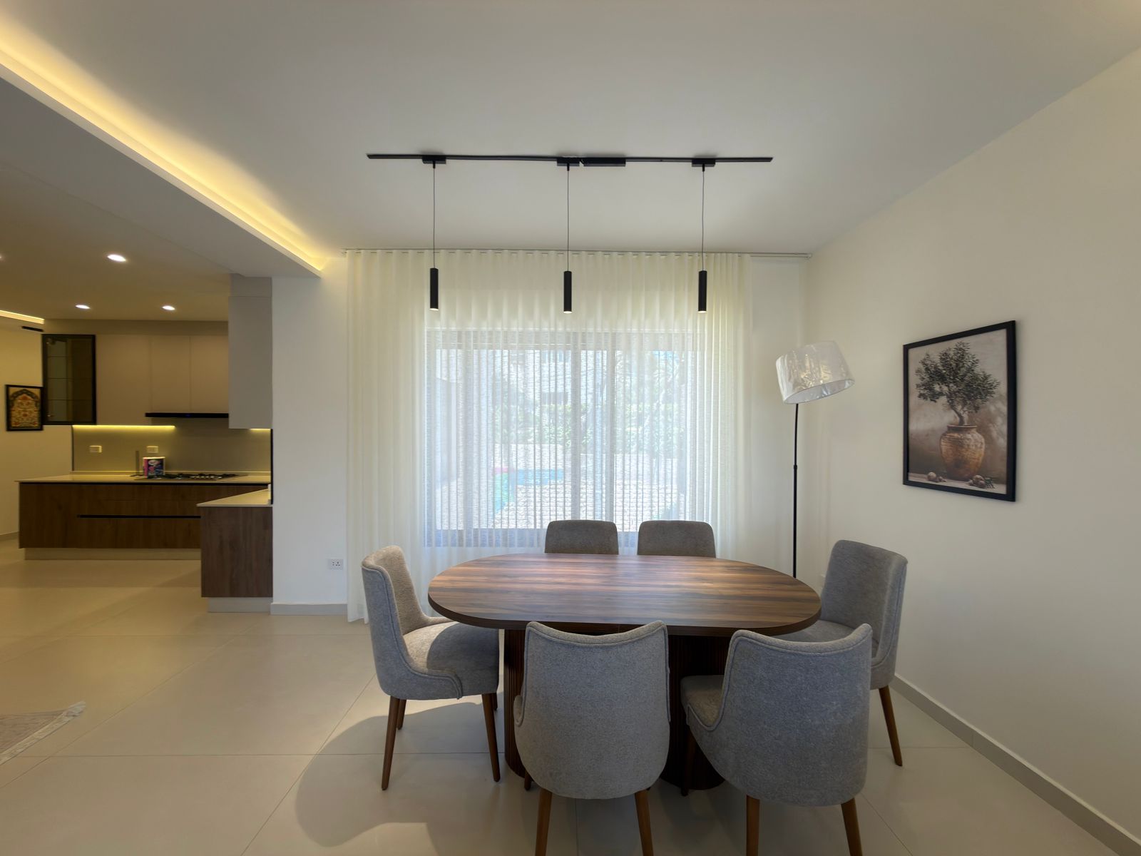 90fff81b-7f52-419f-ba9b-154565cac635 Open-plan dining area with a round wooden table and six gray upholstered chairs, large window with sheer curtains, and pendant lights above.