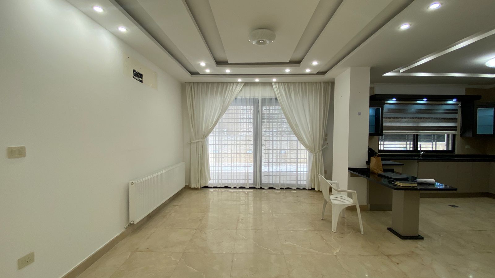 91a430ac-fc4e-457a-9bcd-bdf7783adecc Empty living area with marble floor, white walls, and a radiator on the left; large glass doors with white curtains lead outside, open to a kitchen on the right.