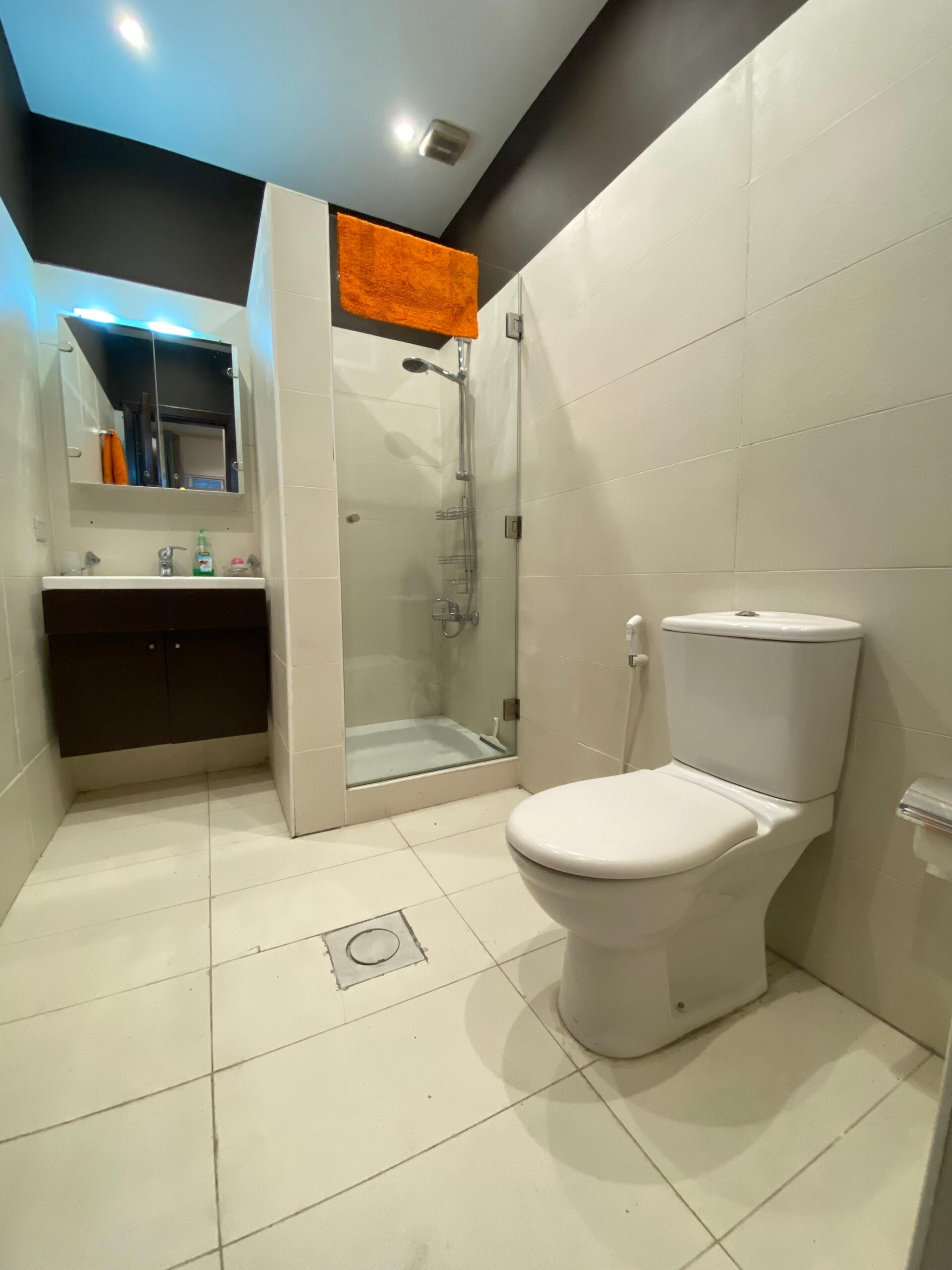 91fd968d-1f58-4863-a75f-f004f0cbd06b Modern bathroom with a glass shower enclosure, white tiles, and a dark vanity; an orange towel hung over the shower wall.
