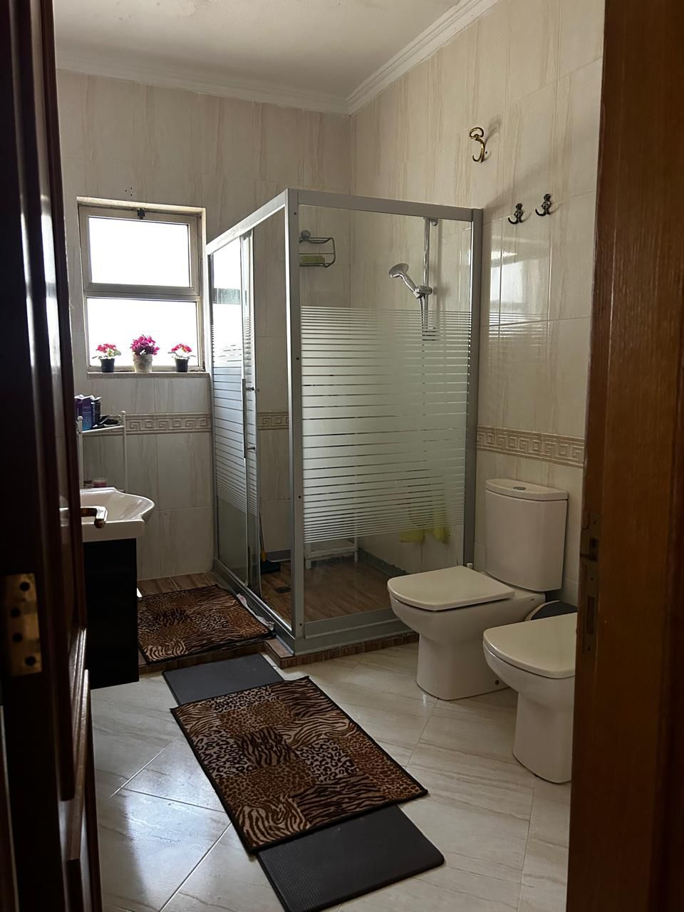 9395f544-d6f0-414c-8674-8383f94721e9 Bright bathroom with a glass-framed shower, toilet and bidet, beige tiled walls, and a window with potted flowers.