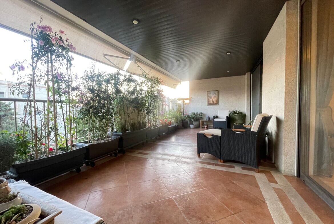 93b4ad63-ce83-48a2-a38d-9c154d40b54a Covered balcony with row of potted plants along the railing and a wicker seating area on a tiled floor.