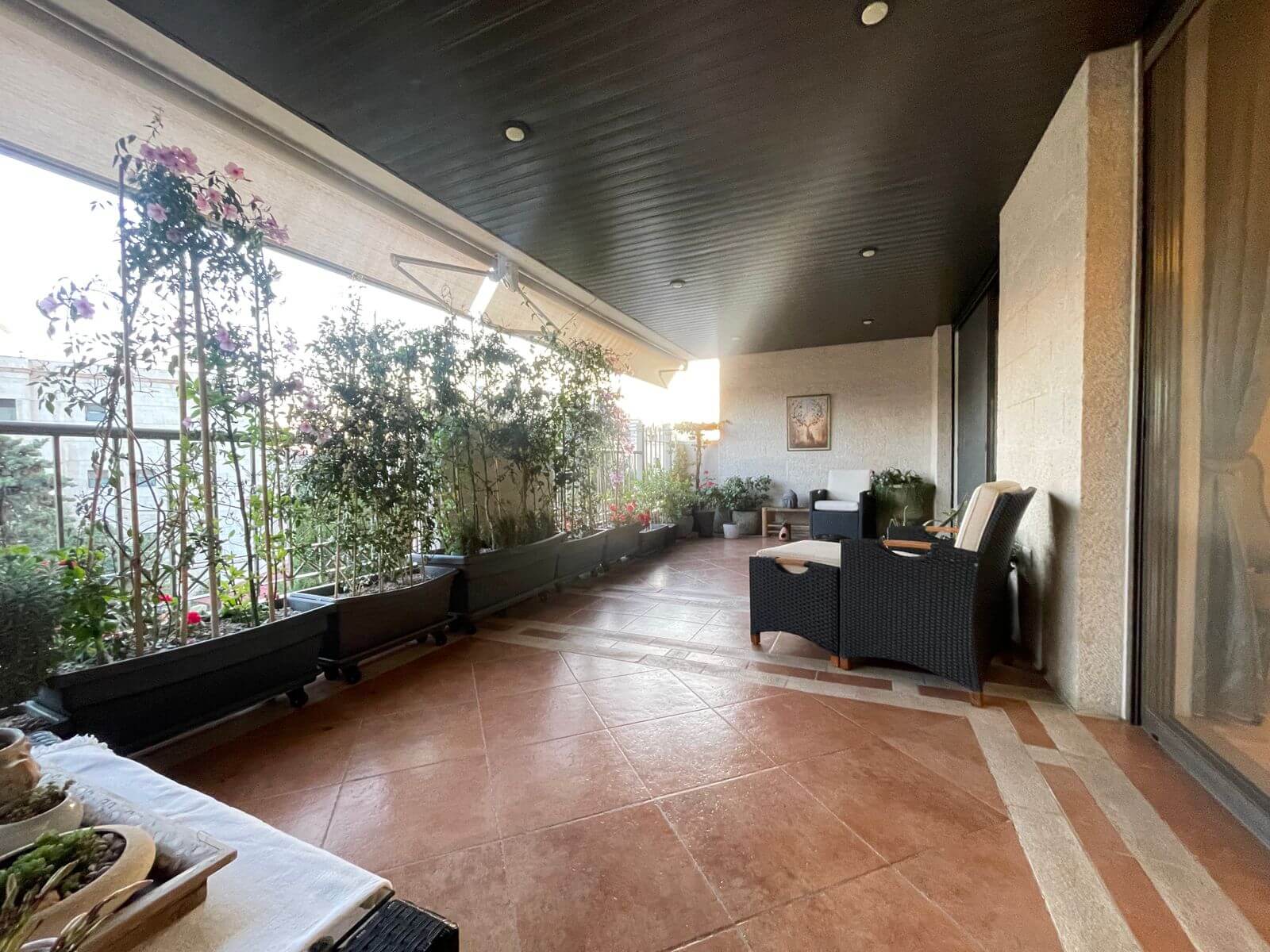 93b4ad63-ce83-48a2-a38d-9c154d40b54a Covered balcony with row of potted plants along the railing and a wicker seating area on a tiled floor.