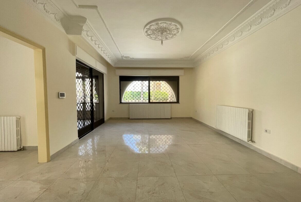 94ef1557-294e-45b4-acba-59f16621e026 Empty living room with marble tile floor, decorative ceiling medallion, wall radiators, and a large arched window with a security grille.