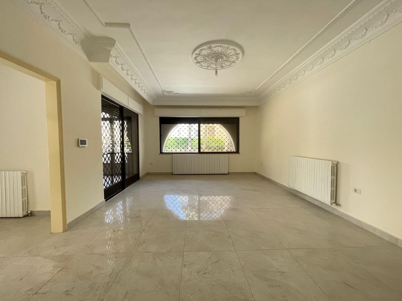94ef1557-294e-45b4-acba-59f16621e026 Empty living room with marble tile floor, decorative ceiling medallion, wall radiators, and a large arched window with a security grille.