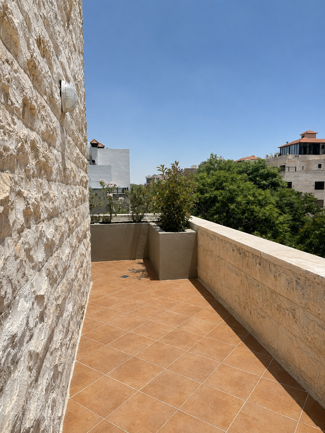 951b7790-4ad6-4e82-8341-4a2dfc311517 Sunny rooftop terrace with terracotta tiles, stone walls, and a rectangular planter with a green shrub, under a clear blue sky.