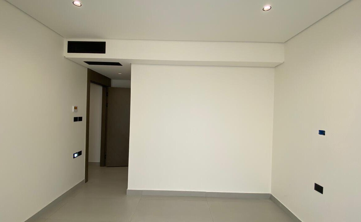 Empty white interior room with a doorway on the left, a partial central wall, and recessed ceiling lights above.