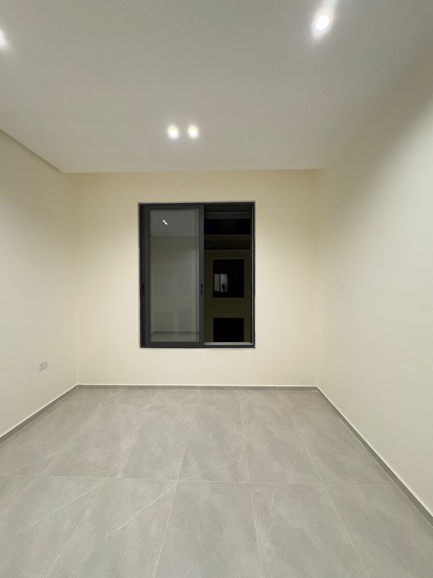 97170b9a-c9bf-41e0-8deb-12ef7d390ccc Empty rectangular room with light gray tiled floor, pale walls, recessed ceiling lights, and a window facing the exterior dark building.