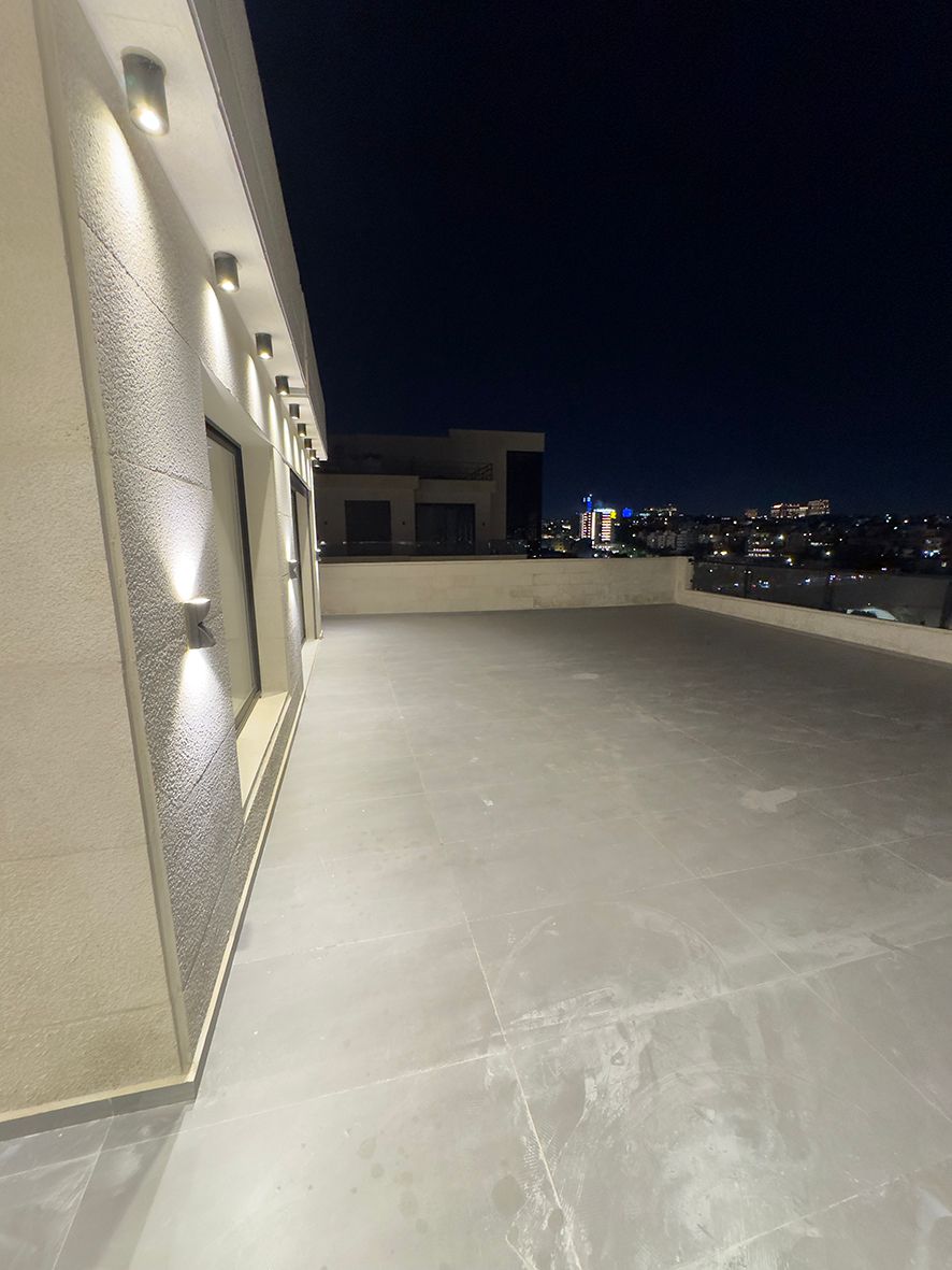976401a1-f1a5-4919-af31-2f14b98aee40 Night view of a spacious rooftop terrace with wall-mounted lights along a beige building on the left and a distant city skyline.