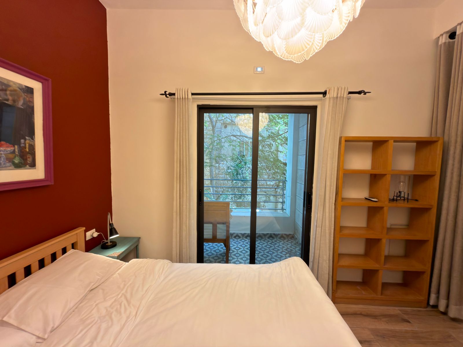 98998dff-0c41-4534-8bad-64fa2bb277bf Bedroom with white bedding and a red accent wall on the left, sliding glass door to a balcony, a wooden bookshelf on the right, and a small bedside lamp on a green table beside the bed.