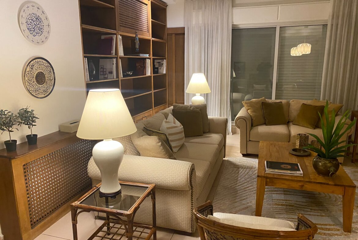 Cozy living room with beige sofas, wooden built-in shelves, two table lamps, and a glass-topped coffee table facing a sliding glass door with sheer curtains.