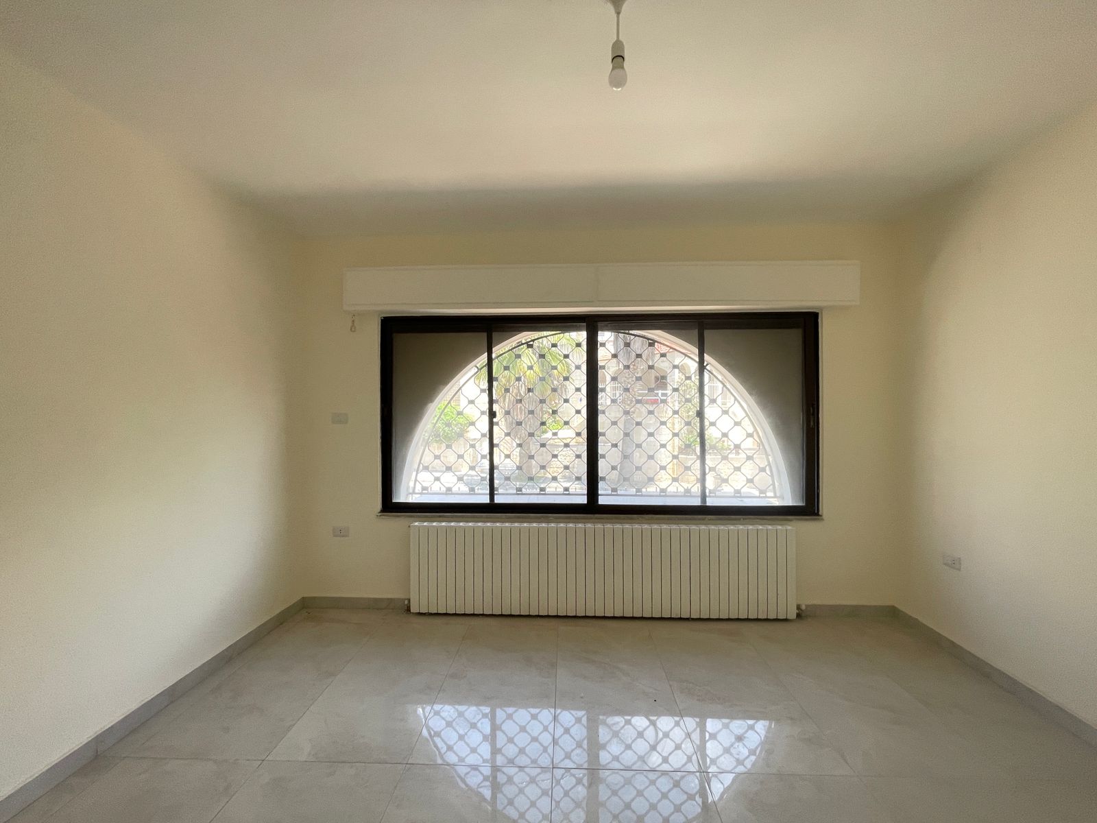 99fee6d9-fd60-40de-ab93-58f46ce1f23f Empty beige room with a large arched window that has a decorative grille and a radiator beneath it.