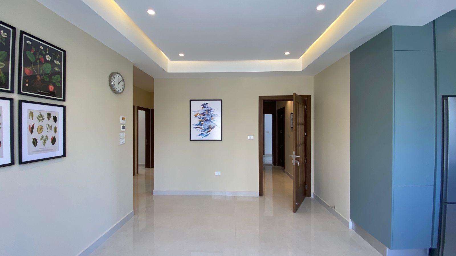9a213168-b8ba-4b7f-81b9-9a5e885c28c0 Bright modern hallway with beige walls, four botanical prints on the left, a round clock, and an open doorway to the right.