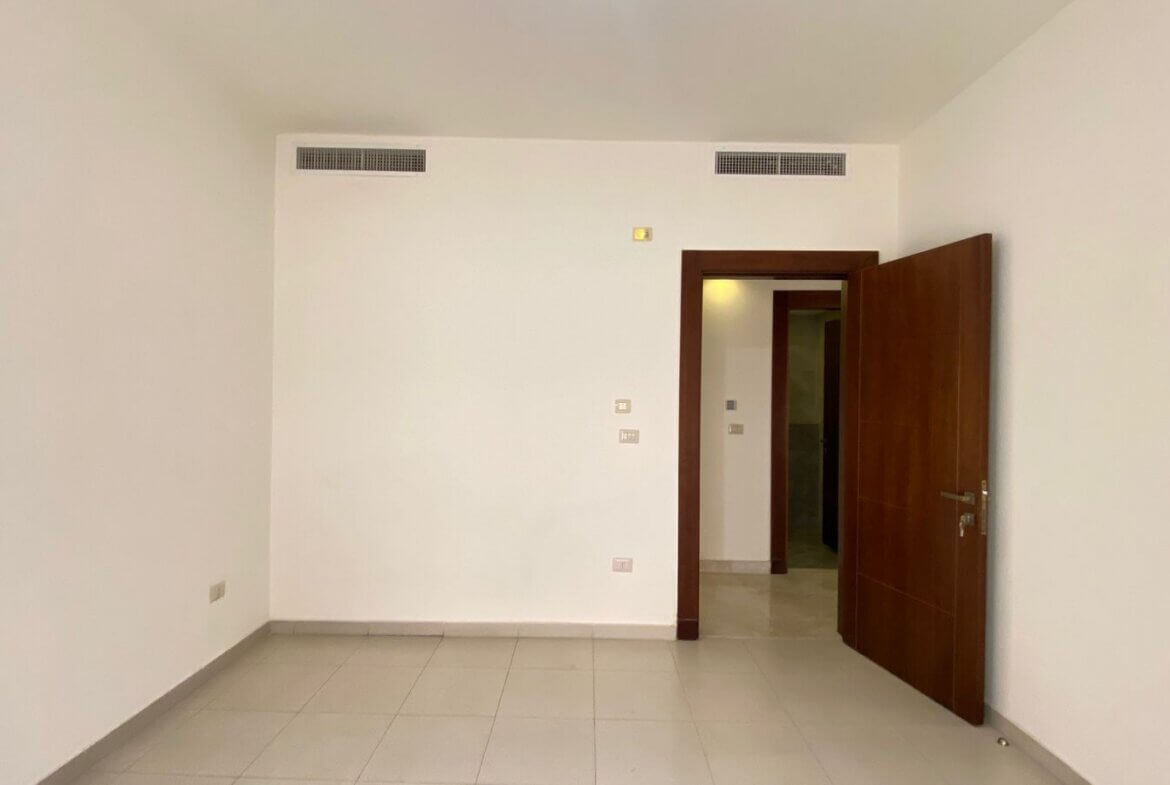 9a49d375-0e57-46a9-868f-1d871bc3e147 Empty room with white walls and light tile floor; an open dark wood door leads to a hallway.