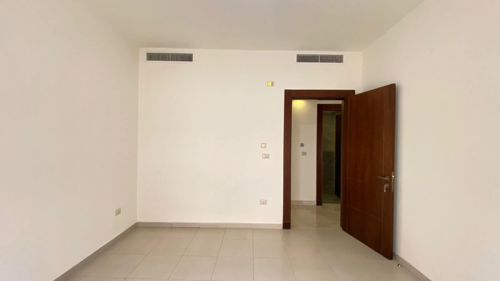 9a49d375-0e57-46a9-868f-1d871bc3e147 Empty room with white walls and light tile floor; an open dark wood door leads to a hallway.
