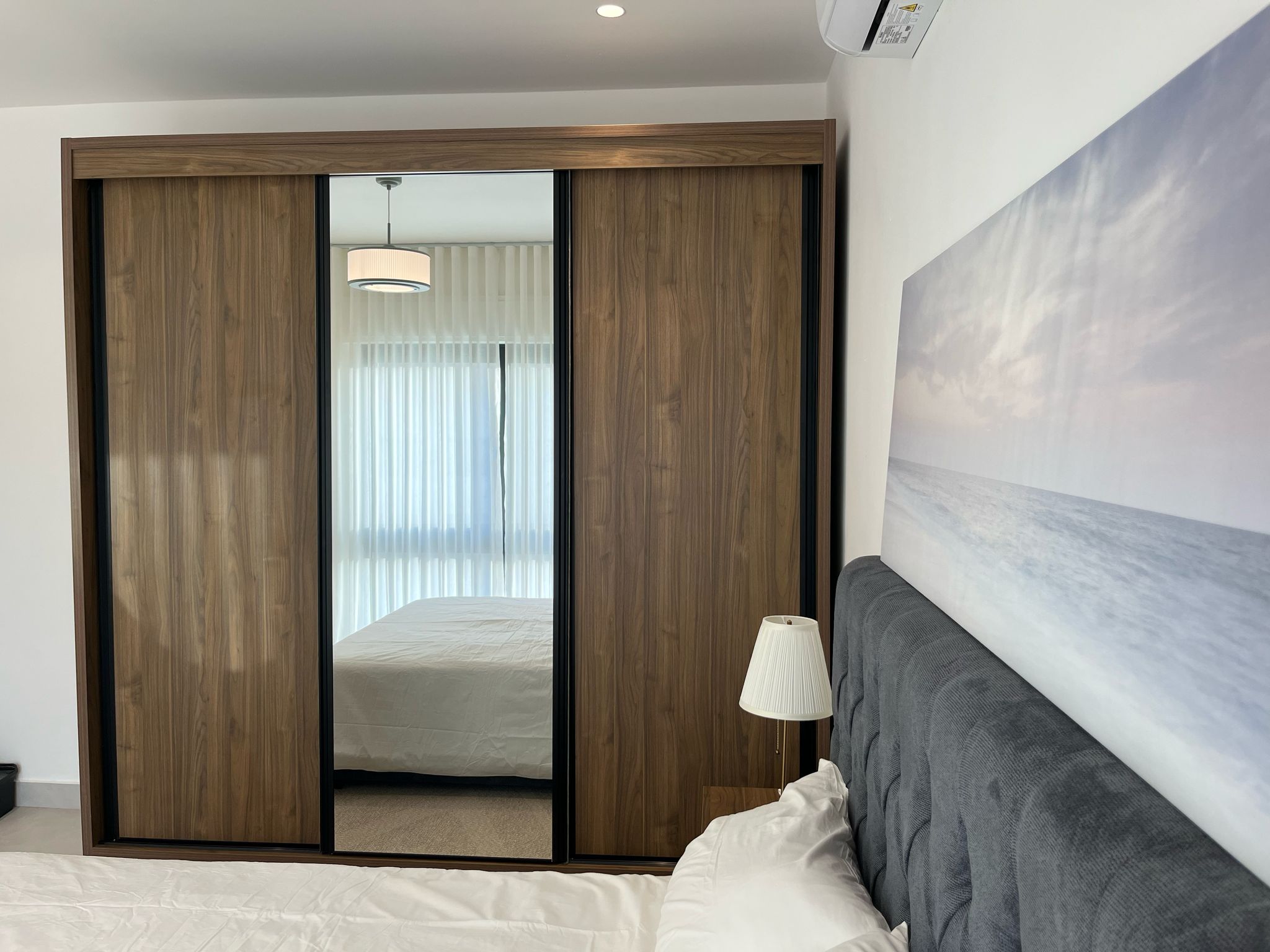 9ac25217-7c8c-43fc-a862-9cc70dc3a0aa Wooden sliding-door wardrobe with a mirrored panel reflecting a bed and pendant light in a modern bedroom.