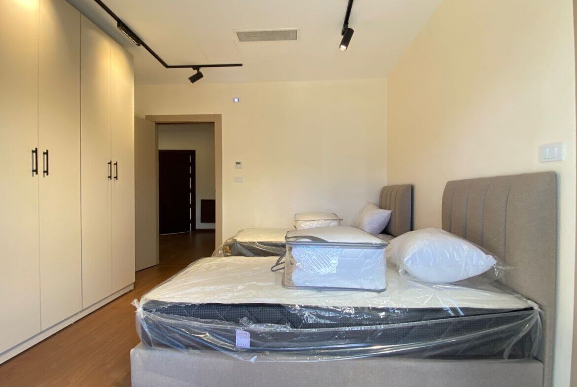 Modern bedroom with two single beds and padded headboards, white walls, and tall built-in wardrobes along the left wall; mattresses still wrapped in plastic for new setup.