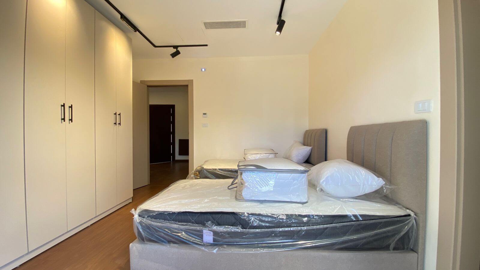 Modern bedroom with two single beds and padded headboards, white walls, and tall built-in wardrobes along the left wall; mattresses still wrapped in plastic for new setup.