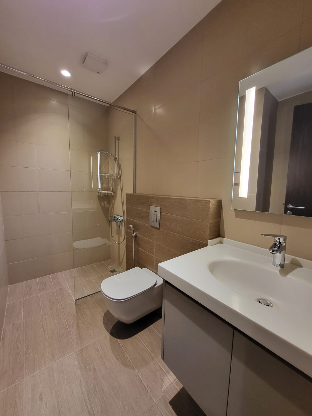 9c446b66-3ee4-40dc-a123-2a4aa68d8776 Modern bathroom with a glass shower enclosure, wall-mounted toilet, white sink, and backlit vertical-mirror on beige tiled walls. Shower area on the left with chrome fixtures and glass partition; toilet and vanity on the right in a contemporary design.