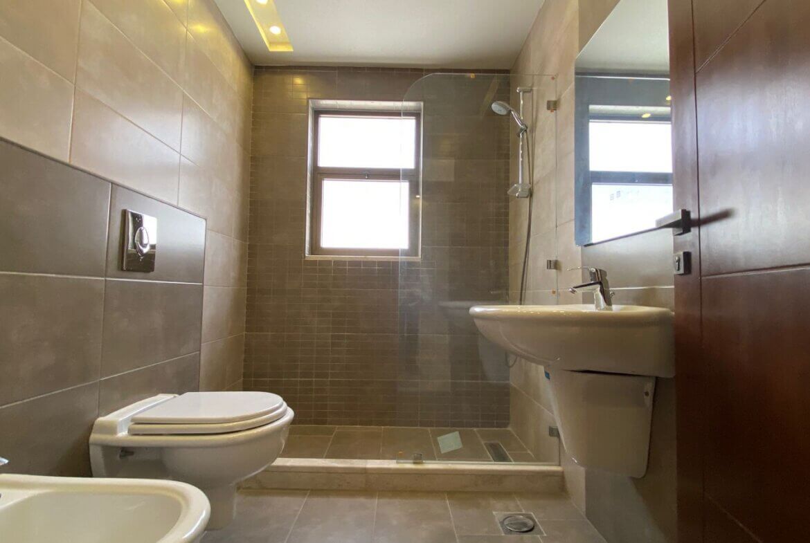 9c873b2f-4b0e-46ec-8b45-4a4518476511 Modern bathroom featuring a wall-mounted toilet, bidet, pedestal sink, and a glass-enclosed shower with grey tile walls and a window in the shower area.