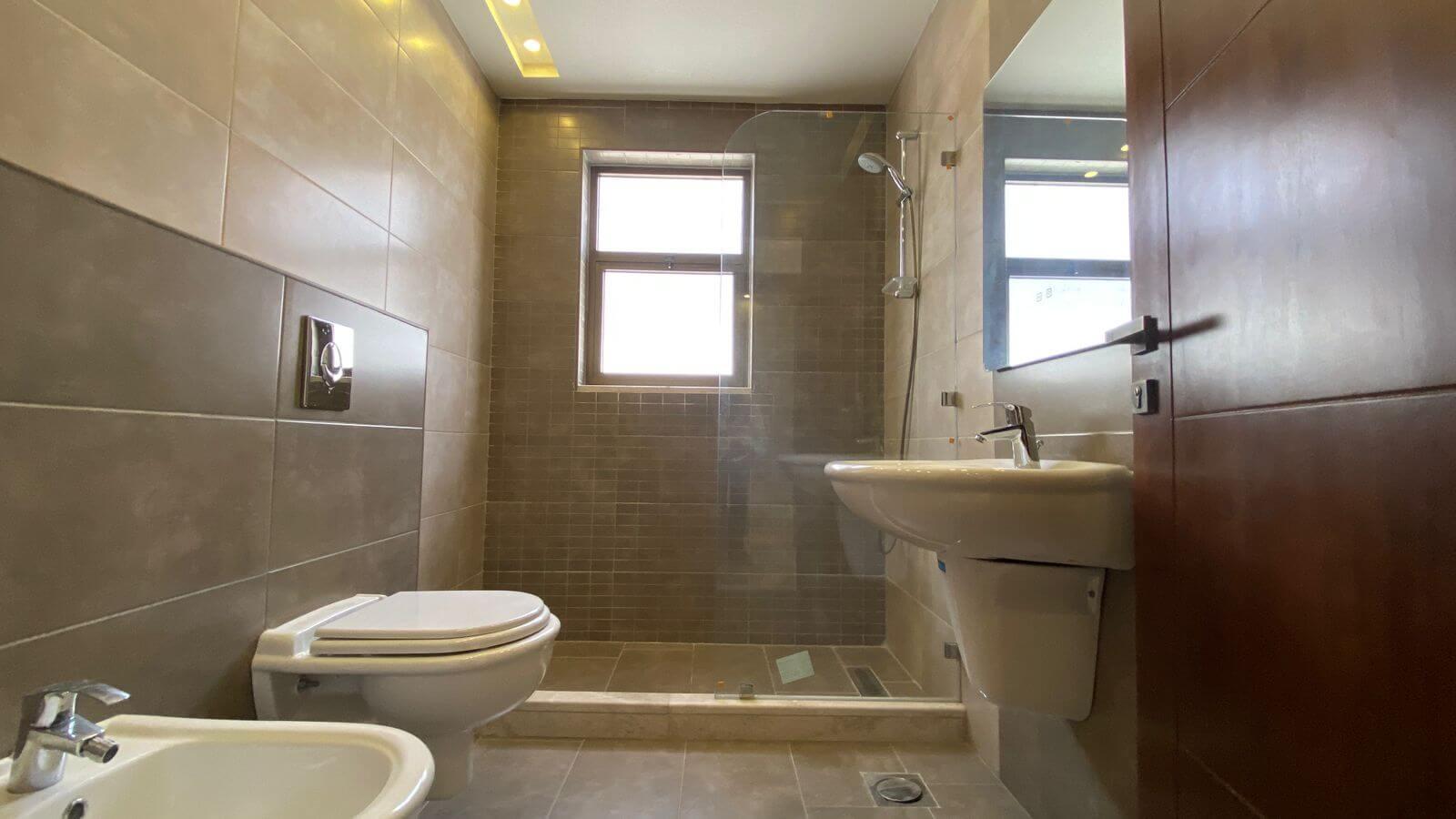 9c873b2f-4b0e-46ec-8b45-4a4518476511 Modern bathroom featuring a wall-mounted toilet, bidet, pedestal sink, and a glass-enclosed shower with grey tile walls and a window in the shower area.