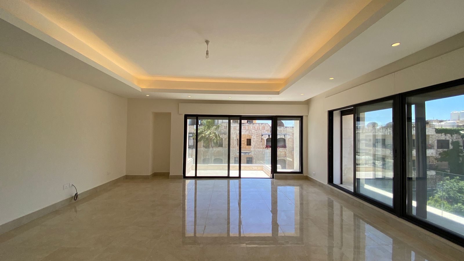 9ca5bd3f-1e73-4267-981b-1b316674e6ac Bright empty living room with large glass sliding doors to a balcony, polished tile floor, and a recessed ceiling with warm backlight.