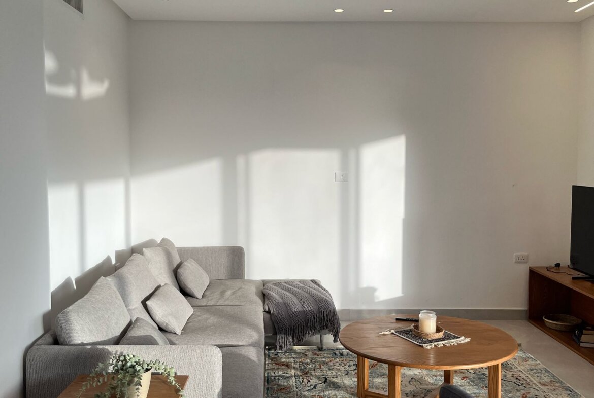 Cozy living room with a light gray sofa, small wooden side table, and a patterned rug bathed in natural light.