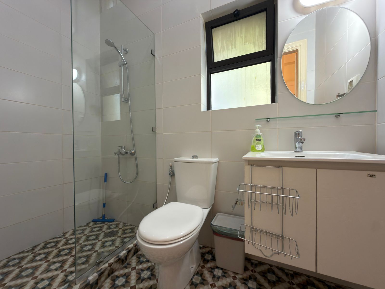 9dd119a0-e956-4880-b1e6-4e27a7bf9e24 Bright bathroom with glass-enclosed shower, toilet, and white vanity with round mirror above and patterned tiled floor