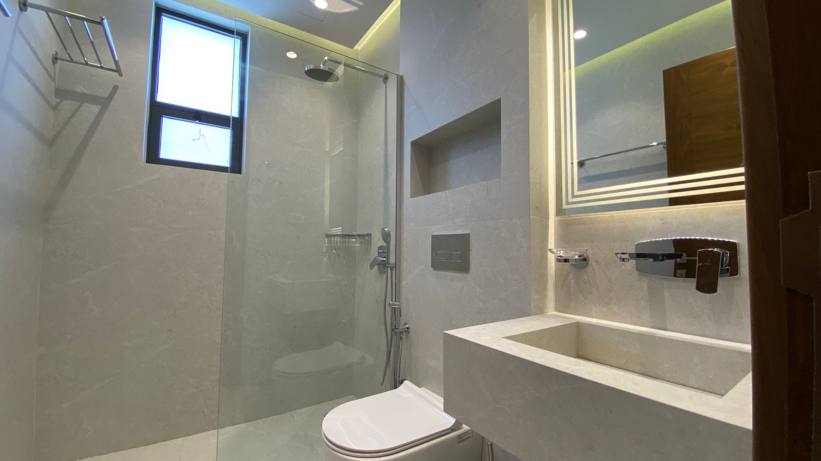 9f5c3dca-c519-47f8-b423-77bd95499770 Modern grey marble bathroom with glass shower enclosure, wall-mounted toilet, and illuminated rectangular mirror above a stone sink.